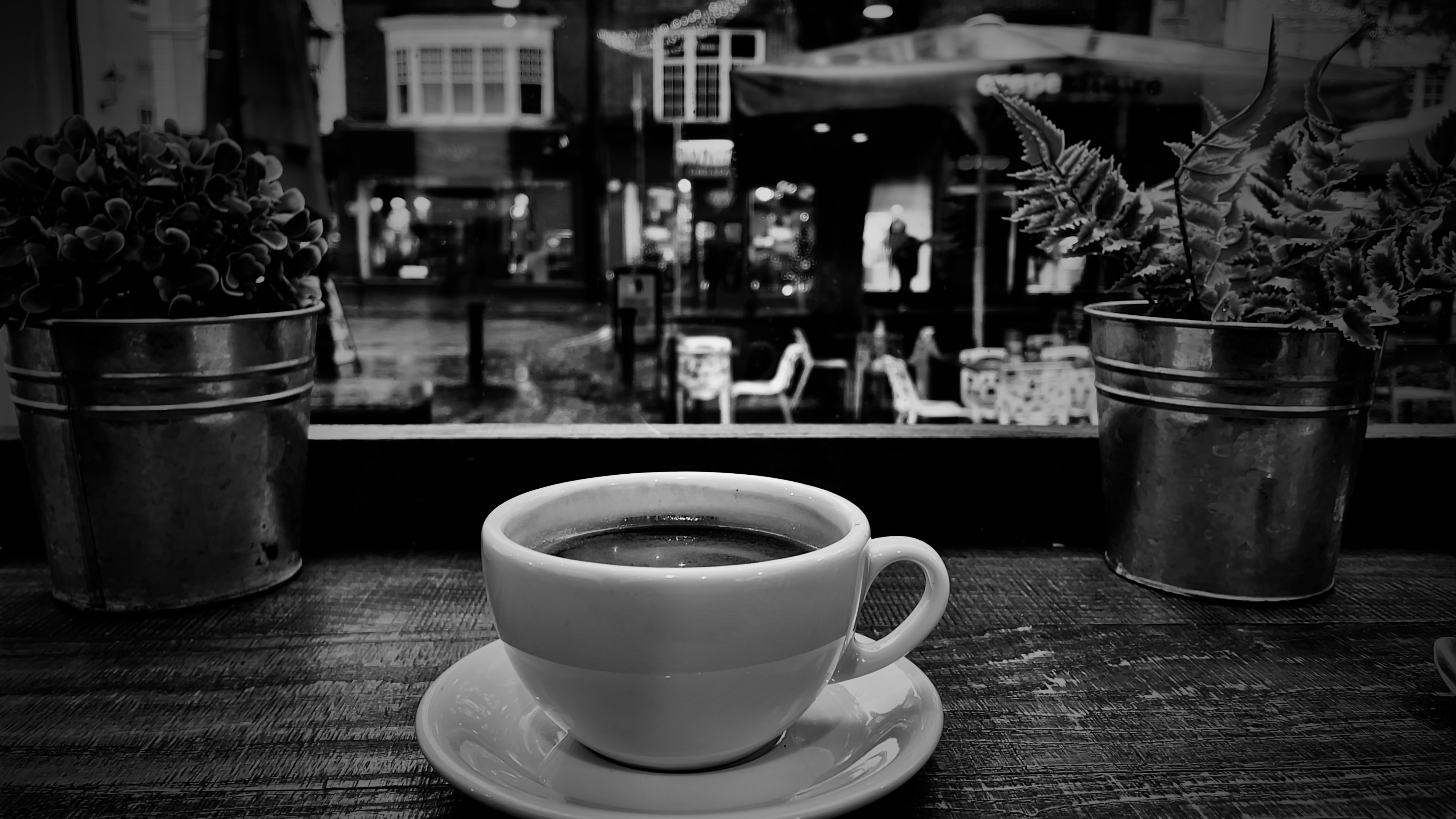 black and white photo of a cup of coffee. beyond the cup is the view out of the window, of a rainy day in brighton